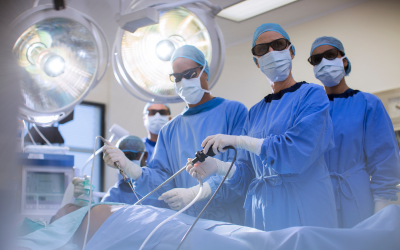 Surgical team in blue scrubs and masks performing an operation under bright overhead lights, using robotic/handheld instruments.