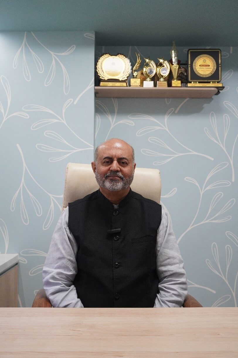 Portrait of a bald man with a gray beard sitting at a desk, facing the camera, with a trophy shelf on his left.
